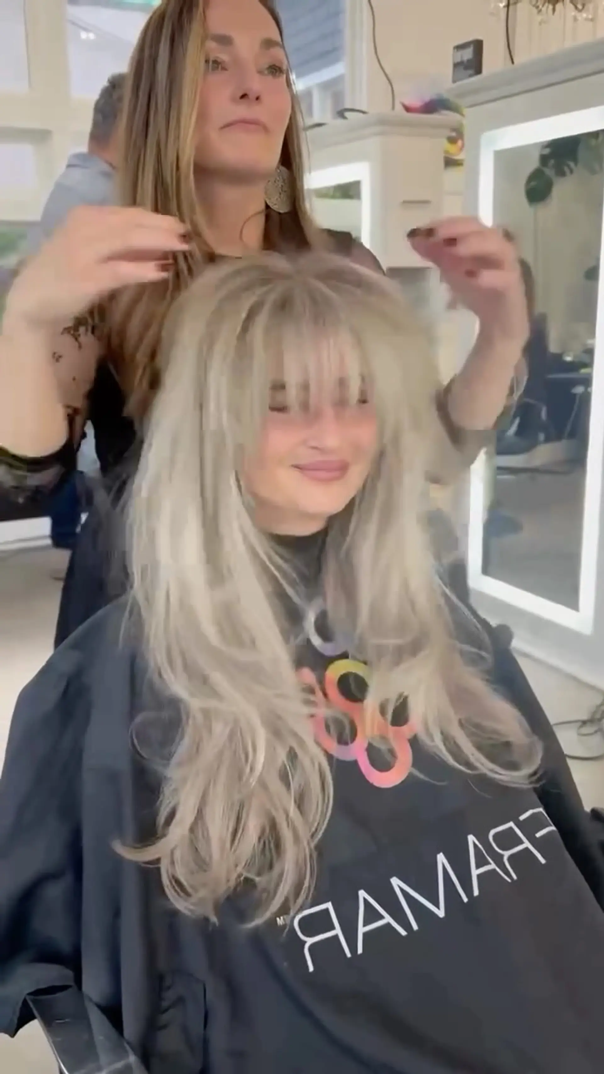 A long ash blonde haircut styled with feathered layers, textured volume, and full eye-skimming bangs. 1
