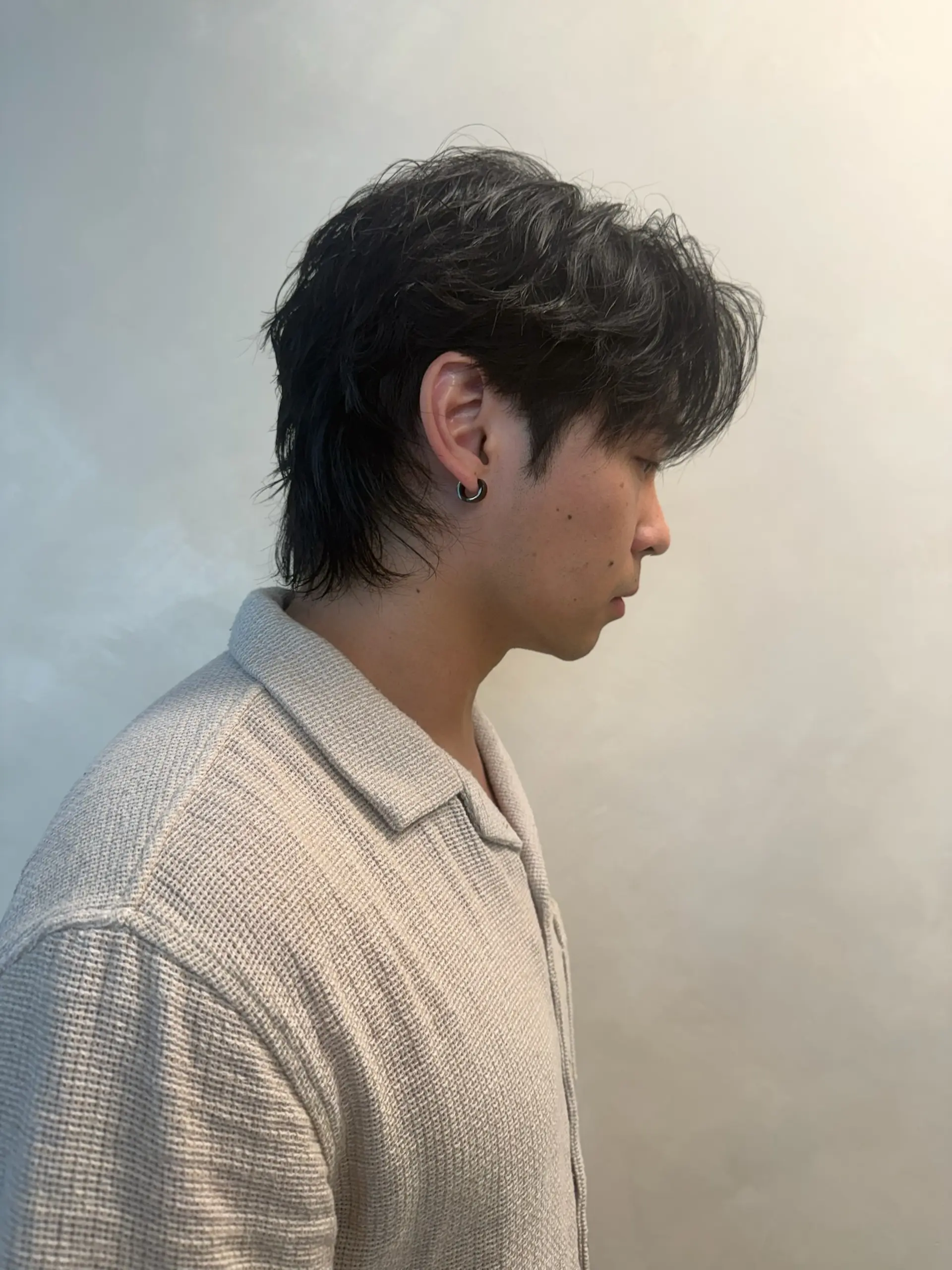 A modern men's haircut featuring soft, textured waves, forward-styled layers, and an elongated length at the nape creating a subtle mullet silhouette. 1