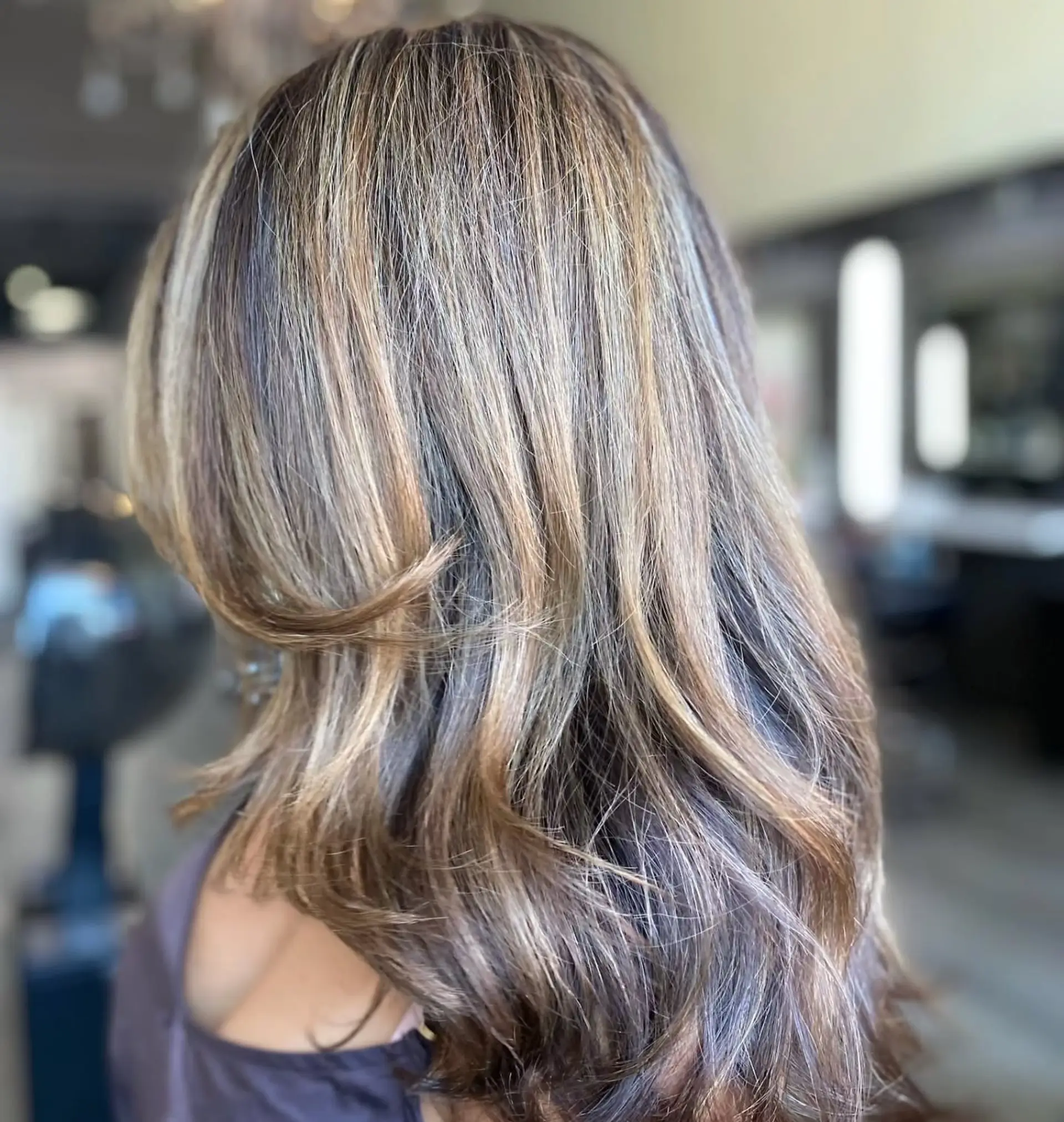 Long layered brunette hair accented with fine dimensional blonde foil highlights and styled into soft, flowing waves. 1