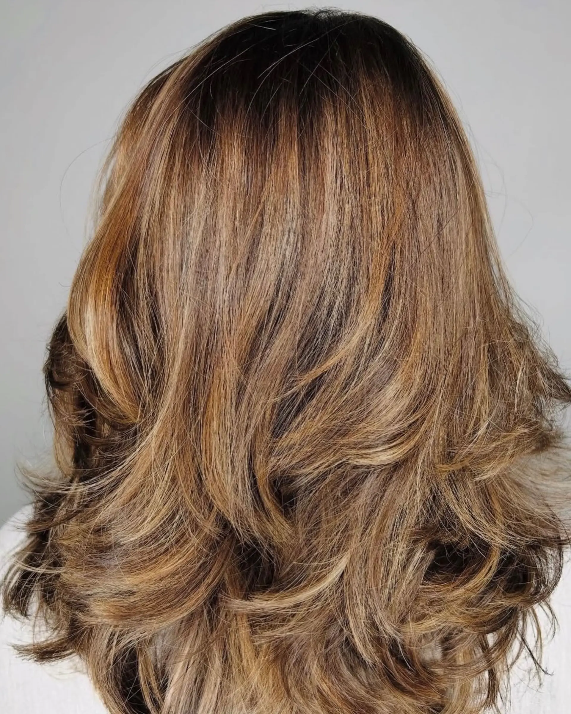 Medium-length textured haircut featuring feathered, flipped layers and blended warm golden-caramel highlights. 1
