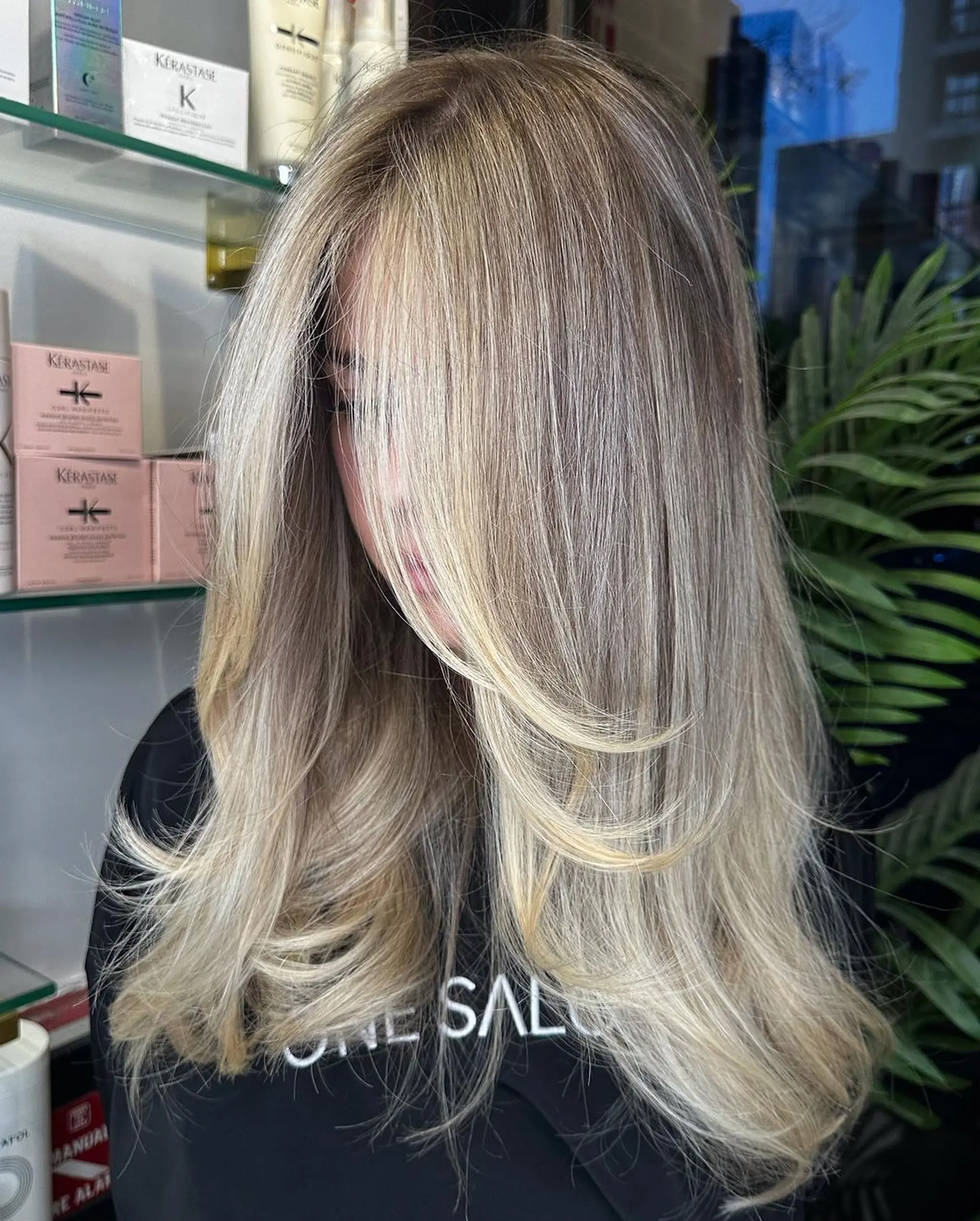 A long, straight hairstyle with blonde, platinum, and light ash brown colors. The hair is layered with a side part, featuring face-framing highlights and natural shine.