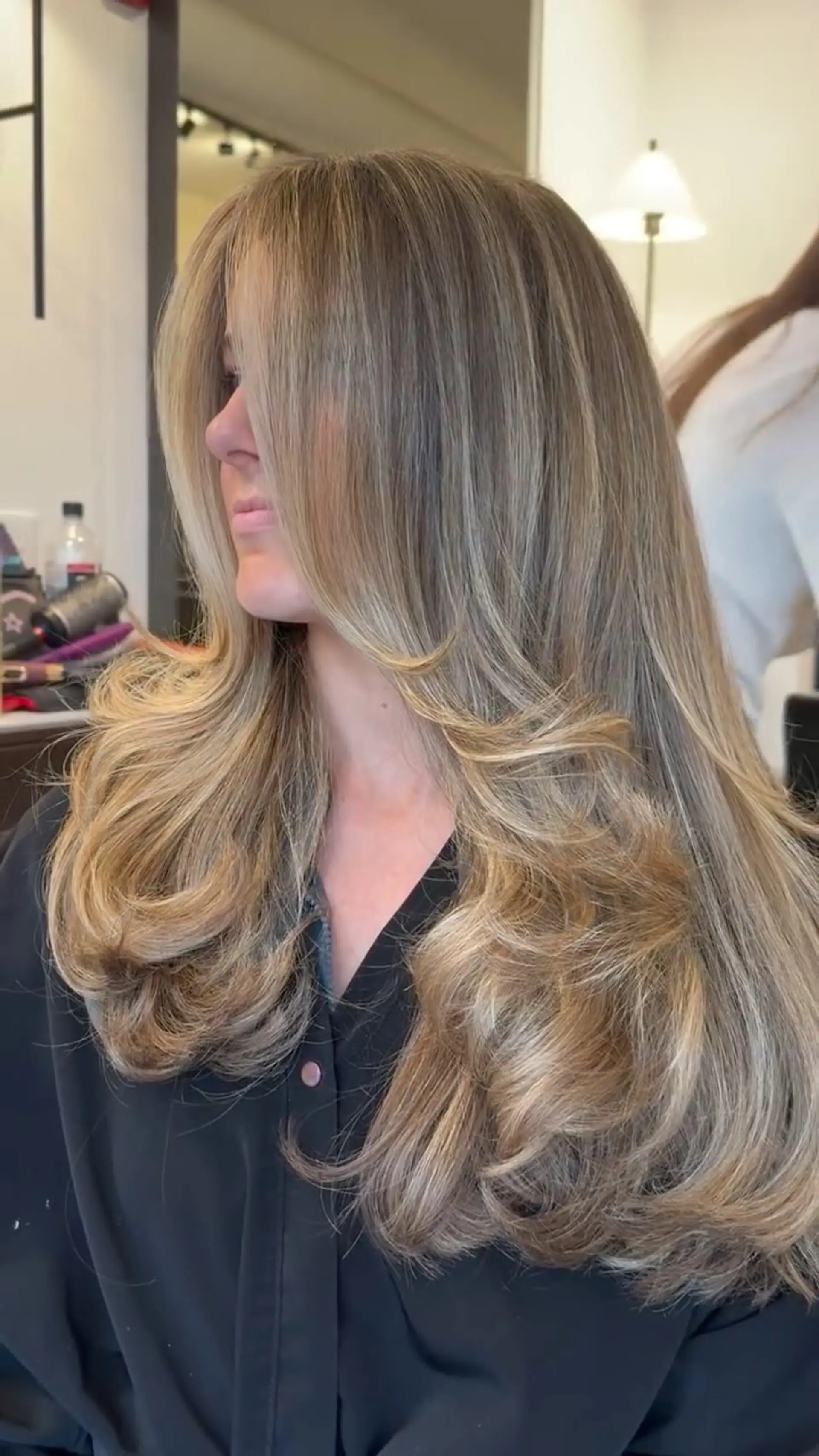 Long multi-layered haircut styled into a full, bouncy blowout with curved flipped ends and face-framing curtain layers, accented by dimensional blonde highlights. 1