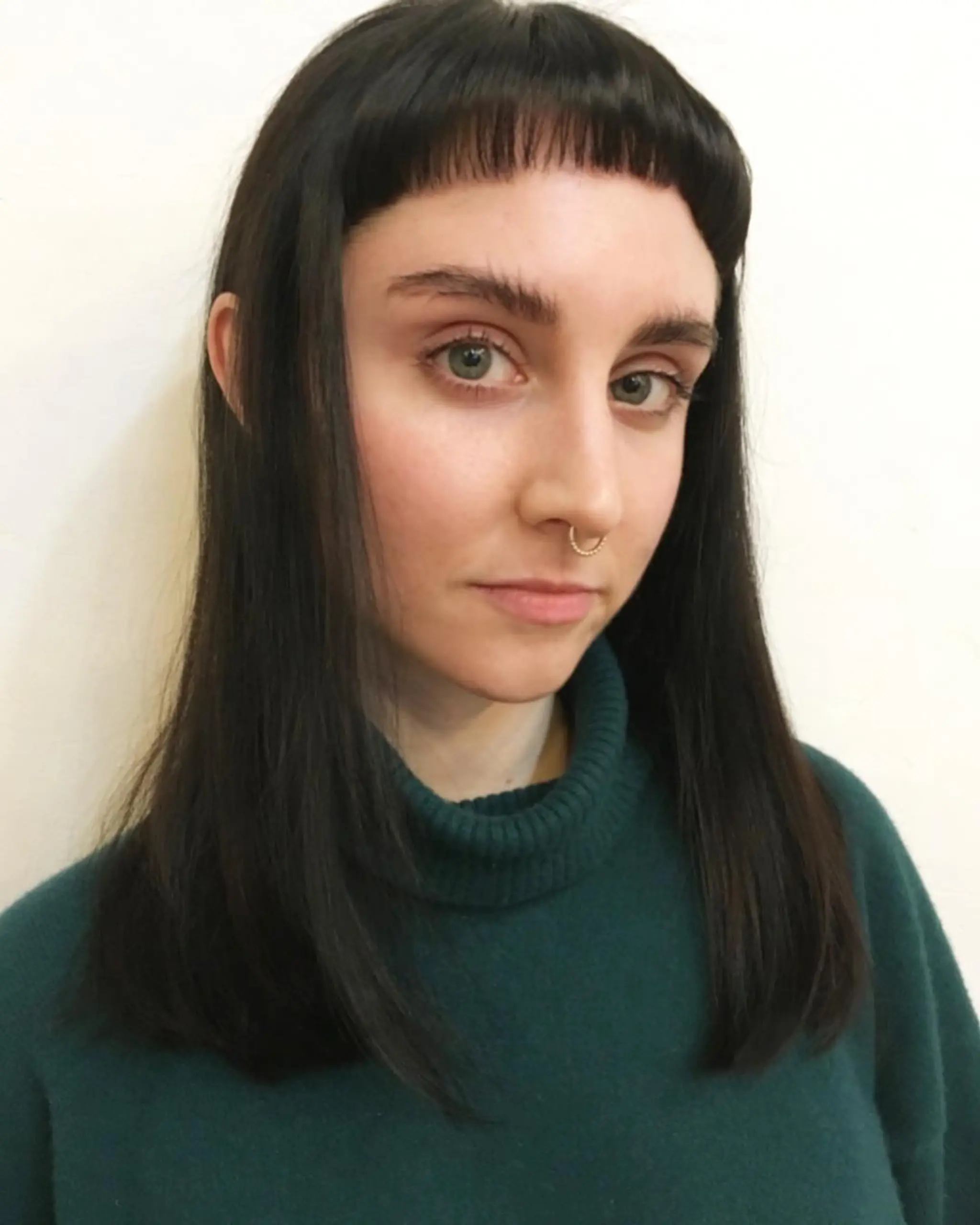 The hair features a bold, blunt fringe that sits high on the forehead, contrasting sharply with the longer, straight black locks that cascade smoothly past the shoulders, creating a chic and edgy look.
