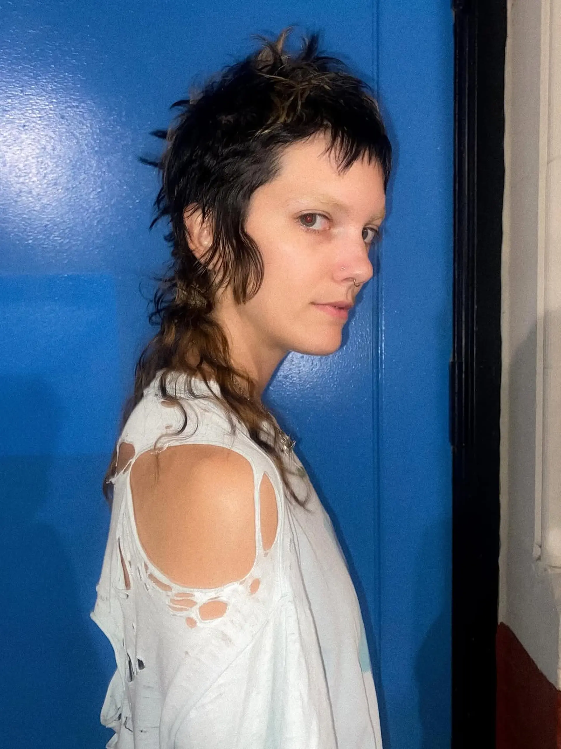 A modern, shaggy mullet cut characterized by short, micro-fringe bangs, heavily textured layers throughout the crown, and a longer, wavy tail in the back. 1