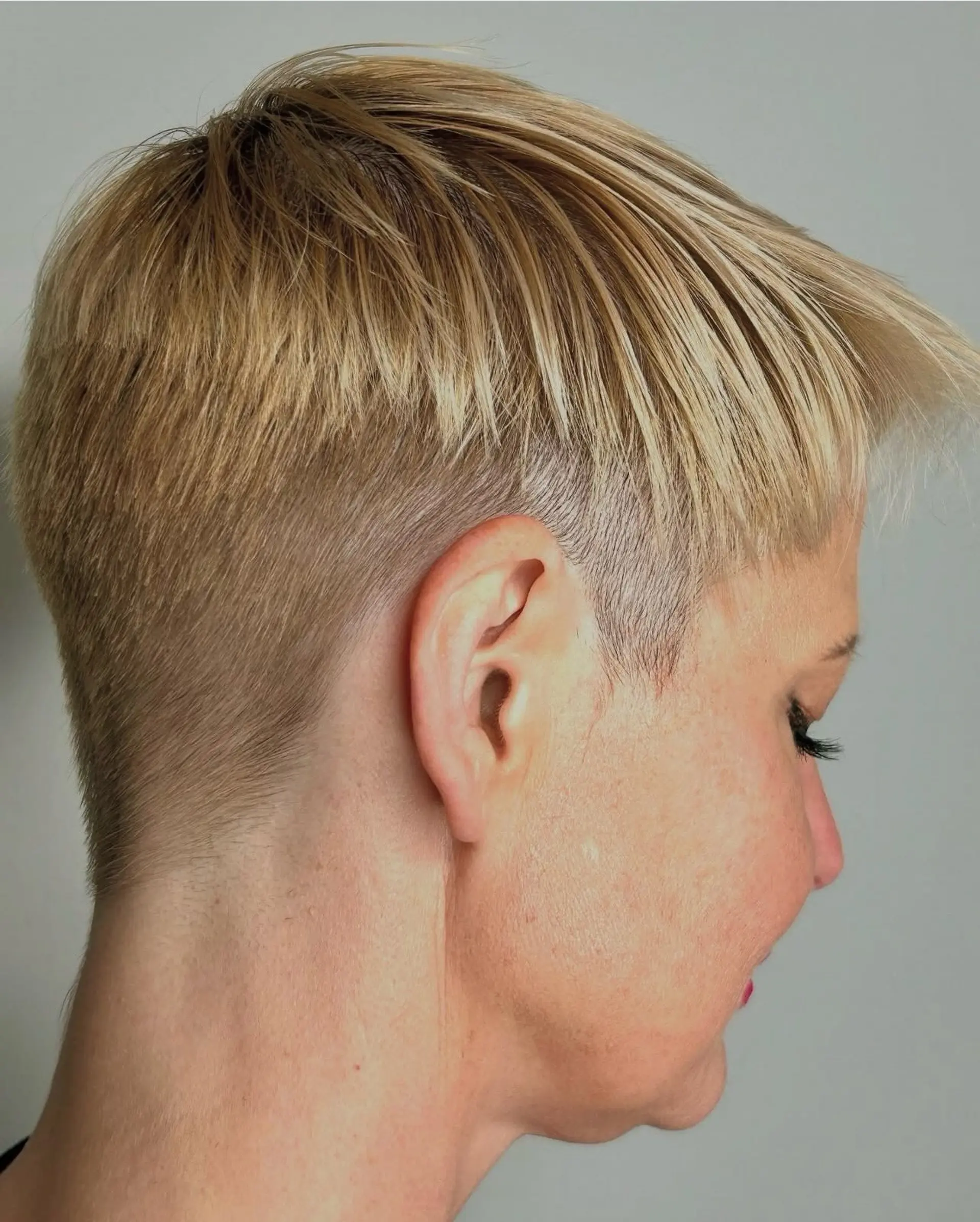 A modern blonde pixie cut featuring a closely buzzed undercut along the sides and nape, topped with textured, forward-styled layers. 1