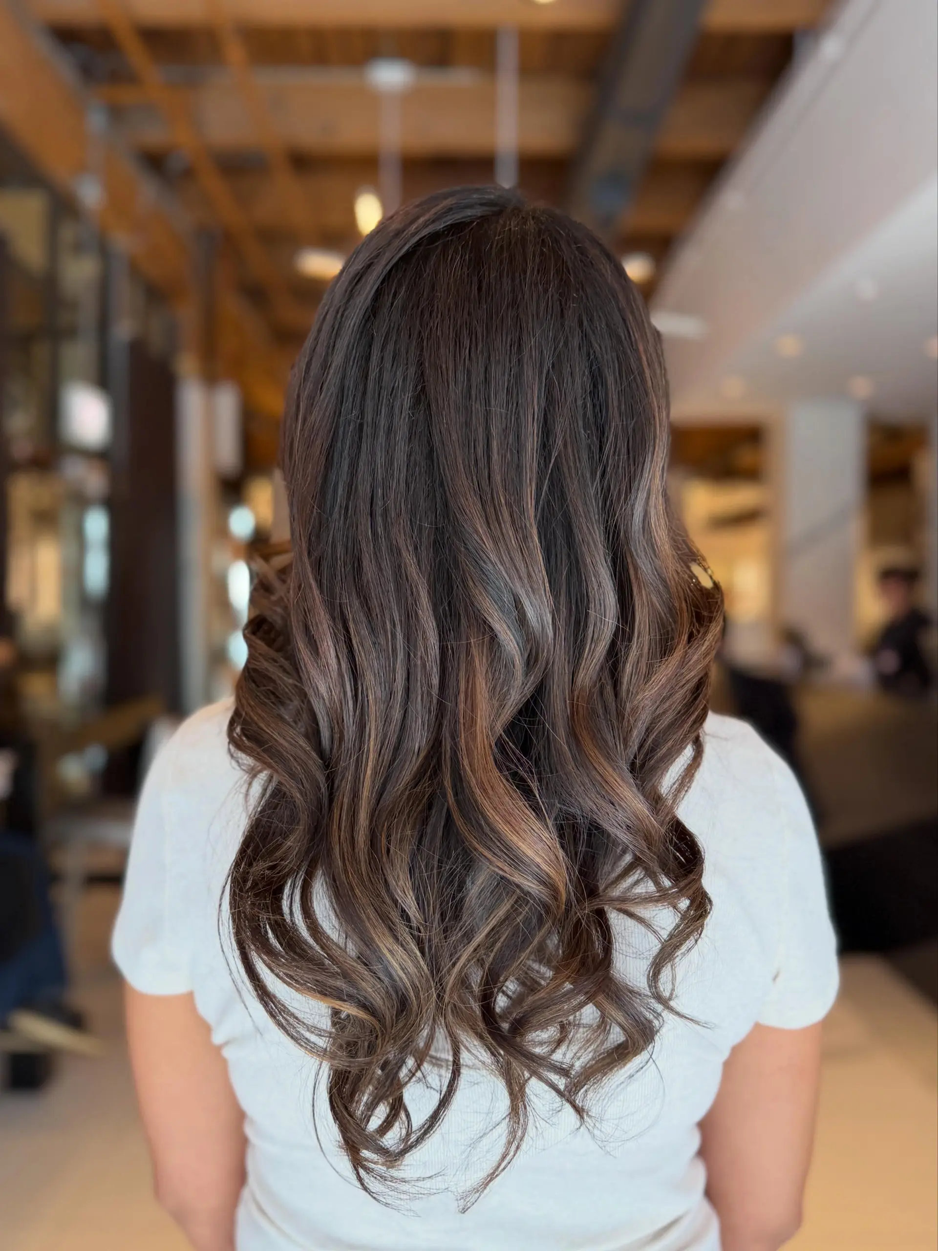 Long chocolate brown hair featuring soft, dimensional caramel balayage highlights styled into polished, bouncy loose waves. 1
