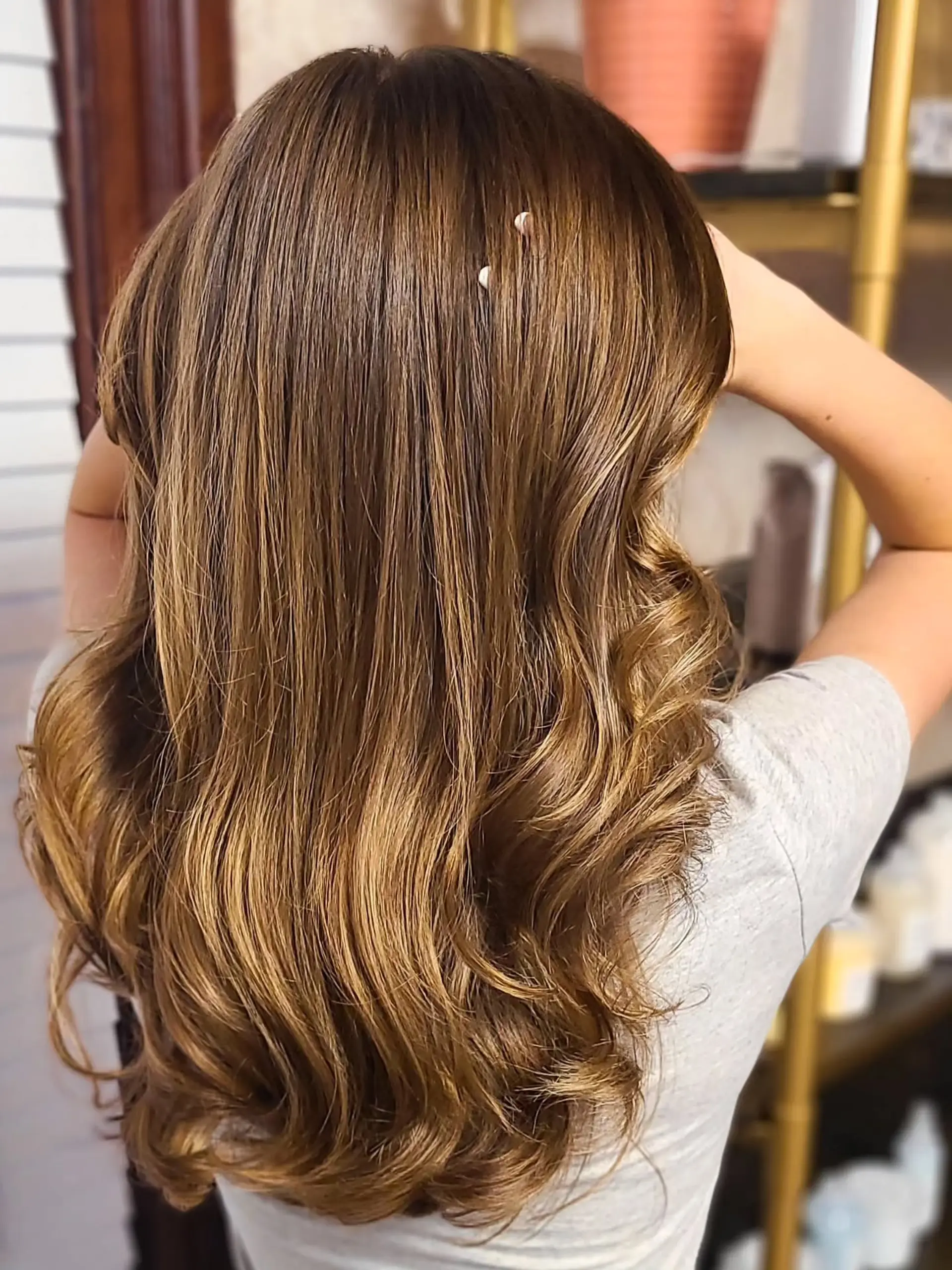 Long, wavy hair with a dimensional golden-brown color, featuring soft, flowing waves that add movement and volume. The color melts seamlessly from a darker root to brighter caramel blonde ends, creating a sun-kissed, lived-in effect. 1