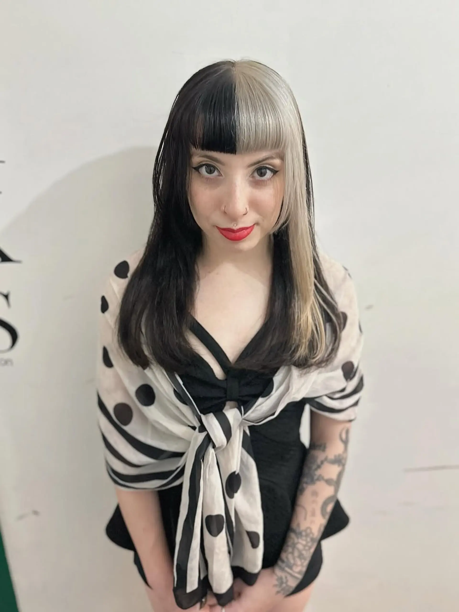 A bold split dye hairstyle featuring half jet black and half platinum blonde hair, styled straight and complemented by sharp, blunt bangs that frame the face. 1